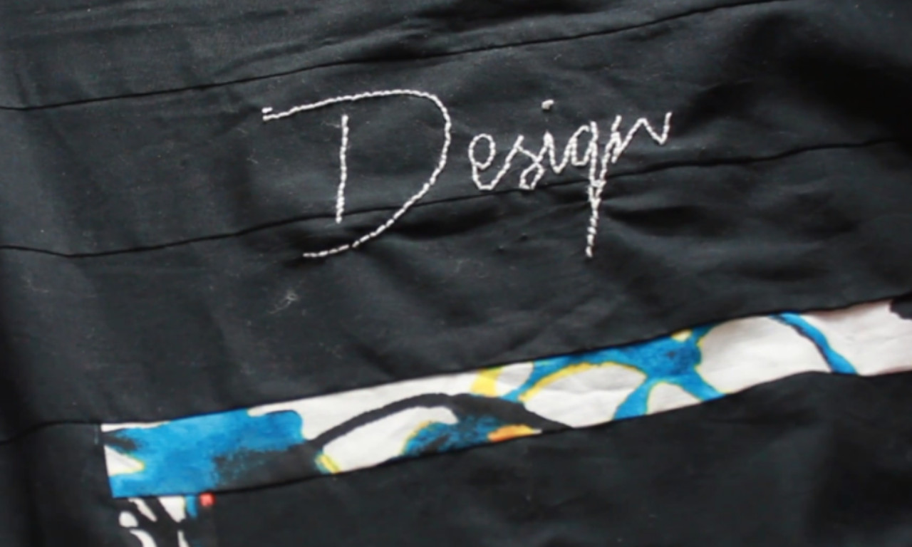 A close-up of a finished embroidery project showing the word "Design" stitched in white cursive font onto black fabric. Below the text is a horizontal strip of colorful, patterned fabric.