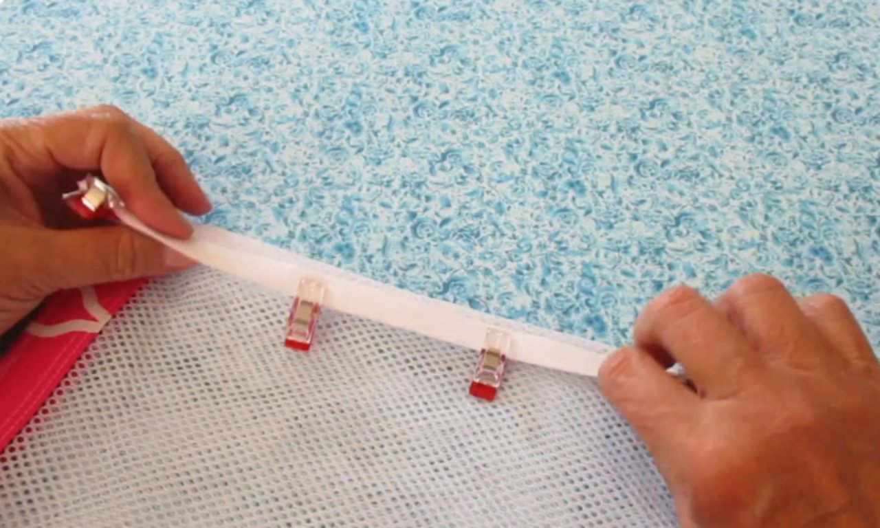 A sewer using red sewing clips to attach white bias tape to the side seam of the mesh laundry bag before sewing.