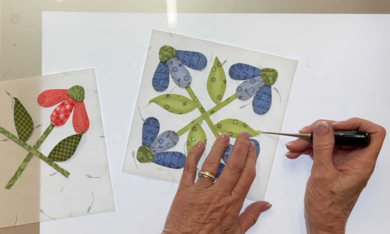 A crafter uses a pointed tool to carefully position a green fabric leaf on a blue floral appliqué quilt block, which is placed on an illuminated lightbox for visibility.