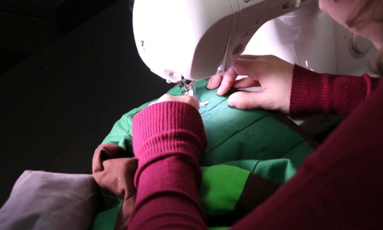 A person carefully guides green fabric under the needle of a sewing machine, stitching a straight line to create a channel for a DIY weighted blanket.
