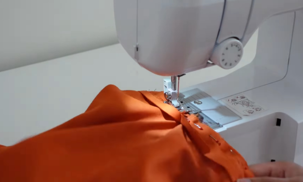 A close-up of a white sewing machine stitching the pinned neckline of the orange boxy top.