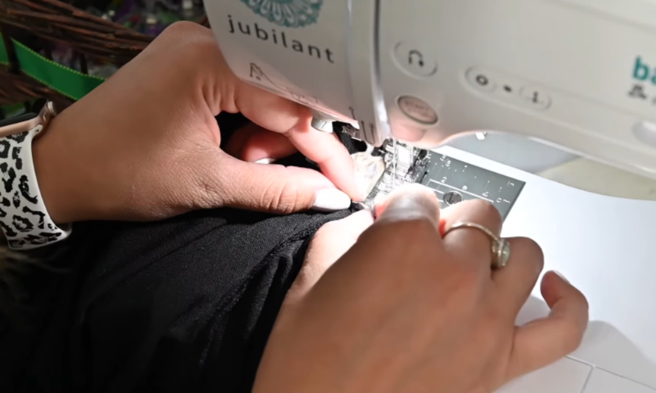 A person using a sewing machine to attach a new side panel to a black t-shirt, carefully guiding the fabric under the needle.