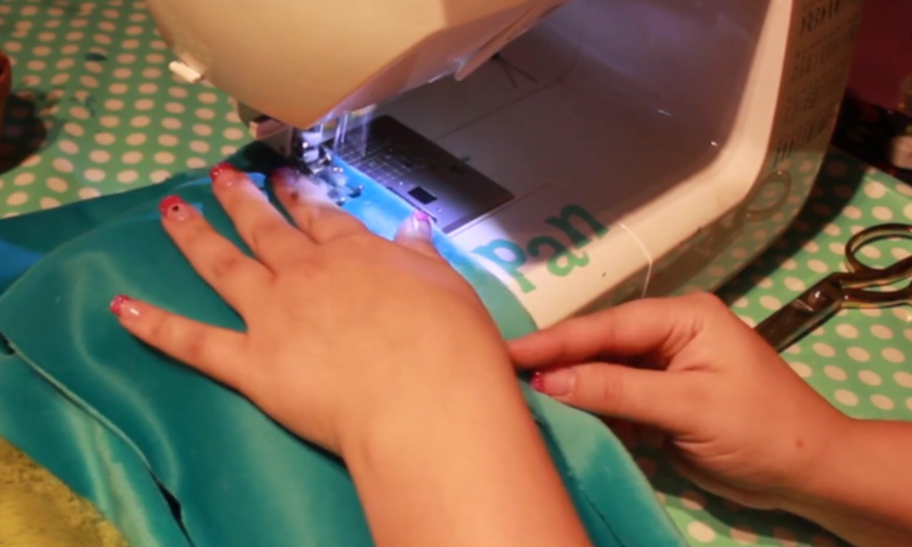 A close-up of a person's hands guiding shiny teal fabric under the needle of a sewing machine.