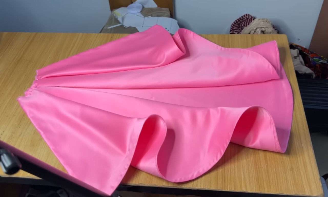 A bright pink satin overskirt, with pleats at the waist and a wavy finished hem, is laid out on a wooden sewing table.
