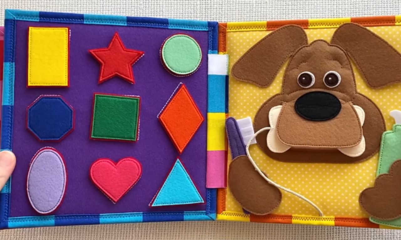 An open fabric quiet book with two educational activity pages. The left page has colorful felt shapes on a purple background for a matching game, while the right page features a friendly felt dog with a toothbrush to practice teeth brushing.