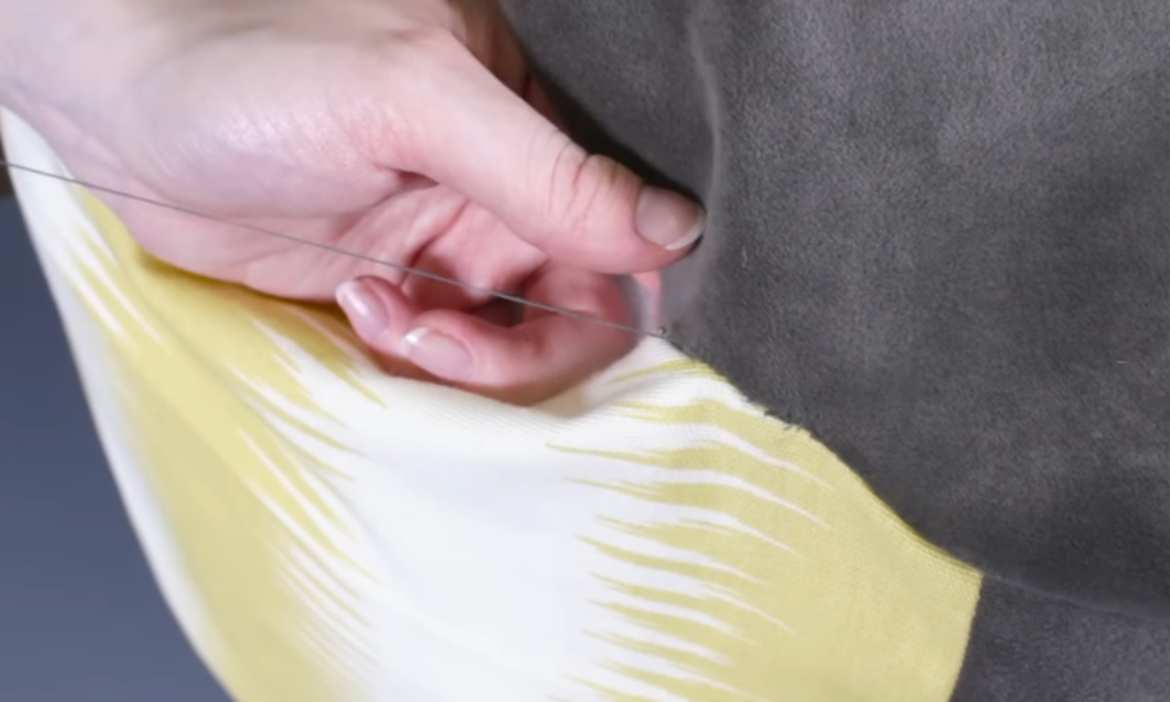 A close-up of a hand using a needle and thread to hand-sew the final seam of a pouf closed, joining a grey fabric panel to a yellow and white patterned one.