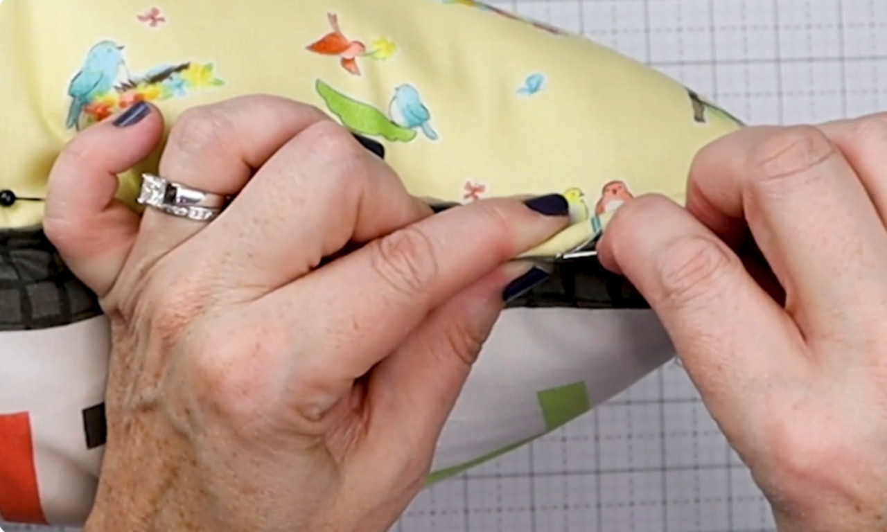 A close-up of a person's hands carefully pushing a pin through the layered fabric of a reading pillow to secure the pieces together before sewing.