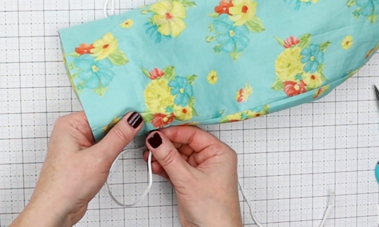 A close-up of a person's hands tying a knot in the white drawstring cord of a handmade blue floral fabric wine bag, set against a gridded cutting mat.