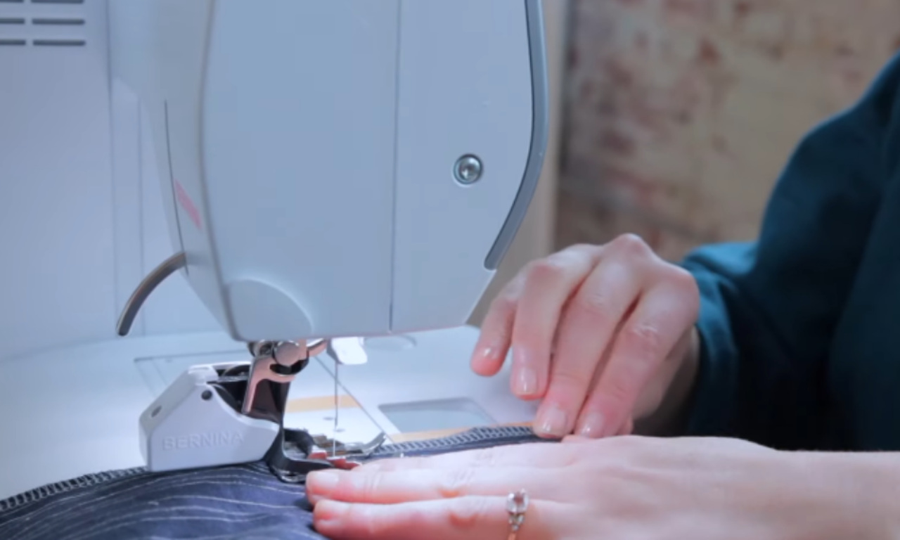 A close-up of a sewer's hands guiding pinstripe fabric under the needle of a sewing machine, stitching a clean seam for the burrito method.