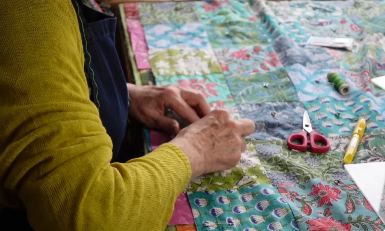 A person wearing a yellow sweater and a blue apron hand-sews a colorful patchwork quilt made of various floral fabrics. Sewing supplies like small red scissors and a spool of green thread are visible on the quilt.