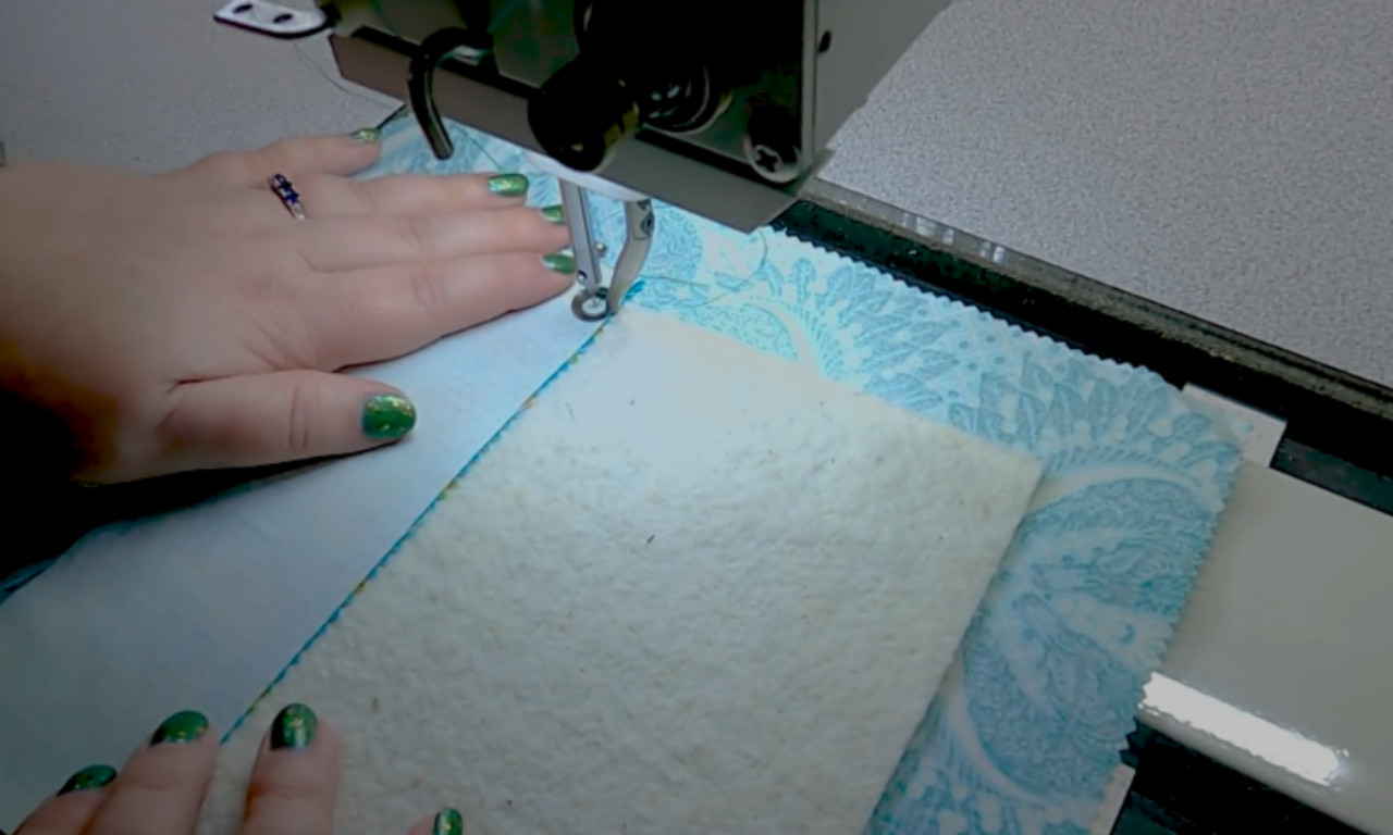 A sewer's hands with green nail polish guiding a light blue fabric scrap onto a square of white batting under the needle of a sewing machine, showing the first step of the quilt-as-you-go technique.