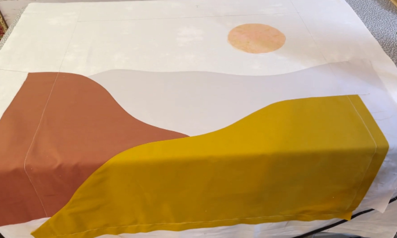A work-in-progress shot of a landscape quilt top being assembled on a table. Pieces of rust, grey, and mustard-yellow fabric are being arranged to form hills against a white background with a peach-colored sun.