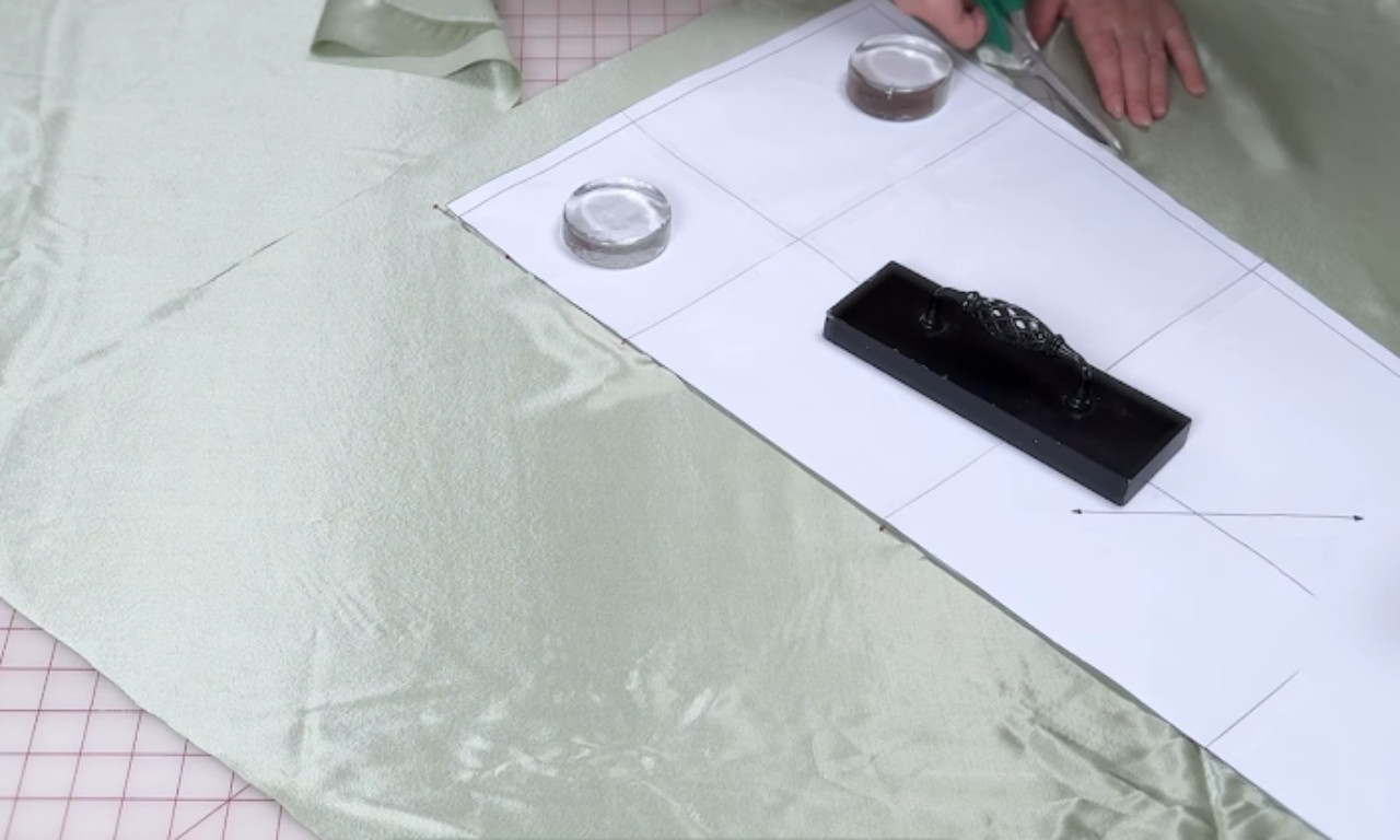 A sewer using scissors to carefully cut sage green fabric around a paper pattern that is held in place with pattern weights on a cutting mat.