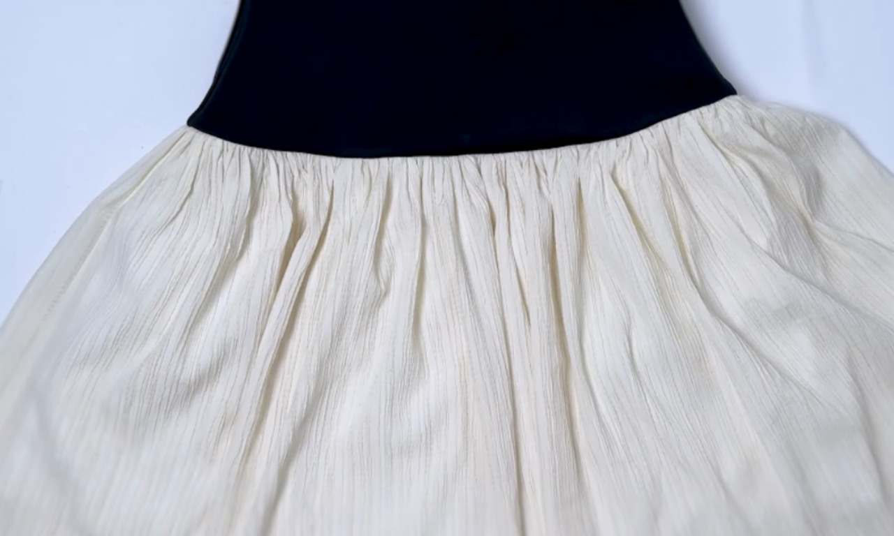 A close-up detail of the drop-waist dress seam, showing the textured and gathered cream skirt neatly attached to the smooth black bodice.