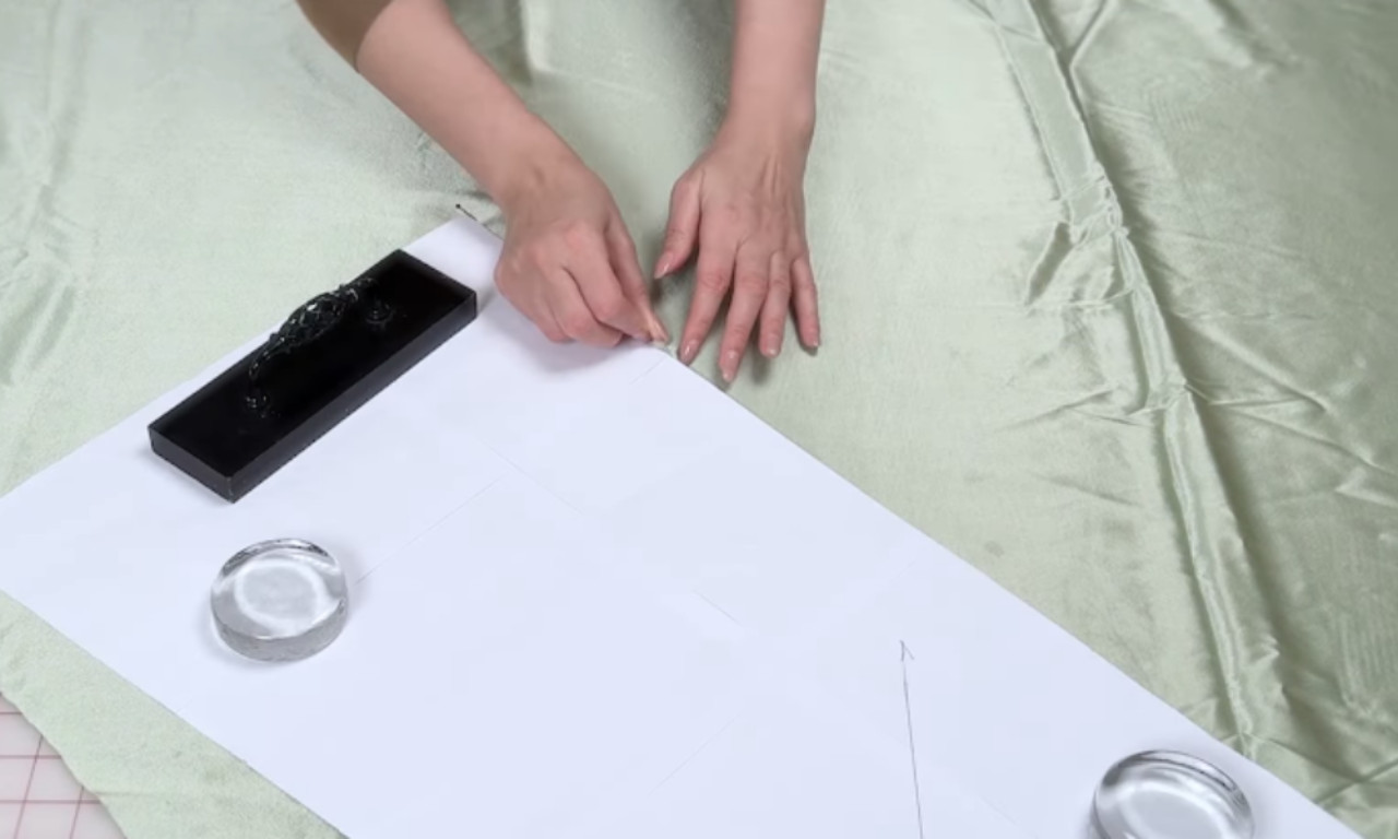 A close-up of a sewer pinning a paper pattern to sage green satin fabric, using pattern weights to hold the slippery material in place before cutting.