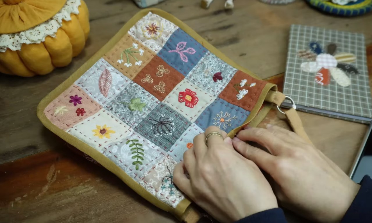 A beautiful, finished patchwork pouch with a mustard-yellow border, showcasing different embroidery on patchwork designs like flowers and leaves on each square.