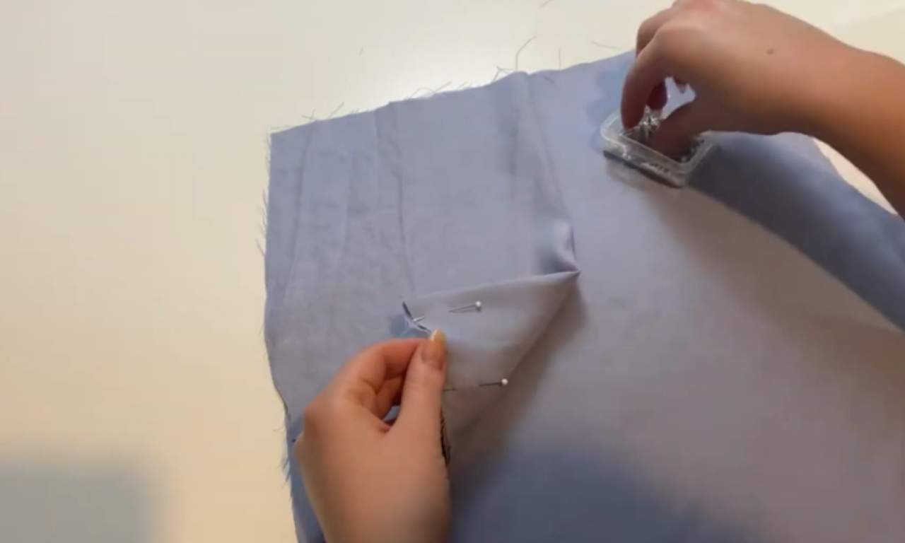 A close-up of a sewer's hands pinning a fold into a piece of light-blue fabric, demonstrating a technique to adjust the fit of pants.