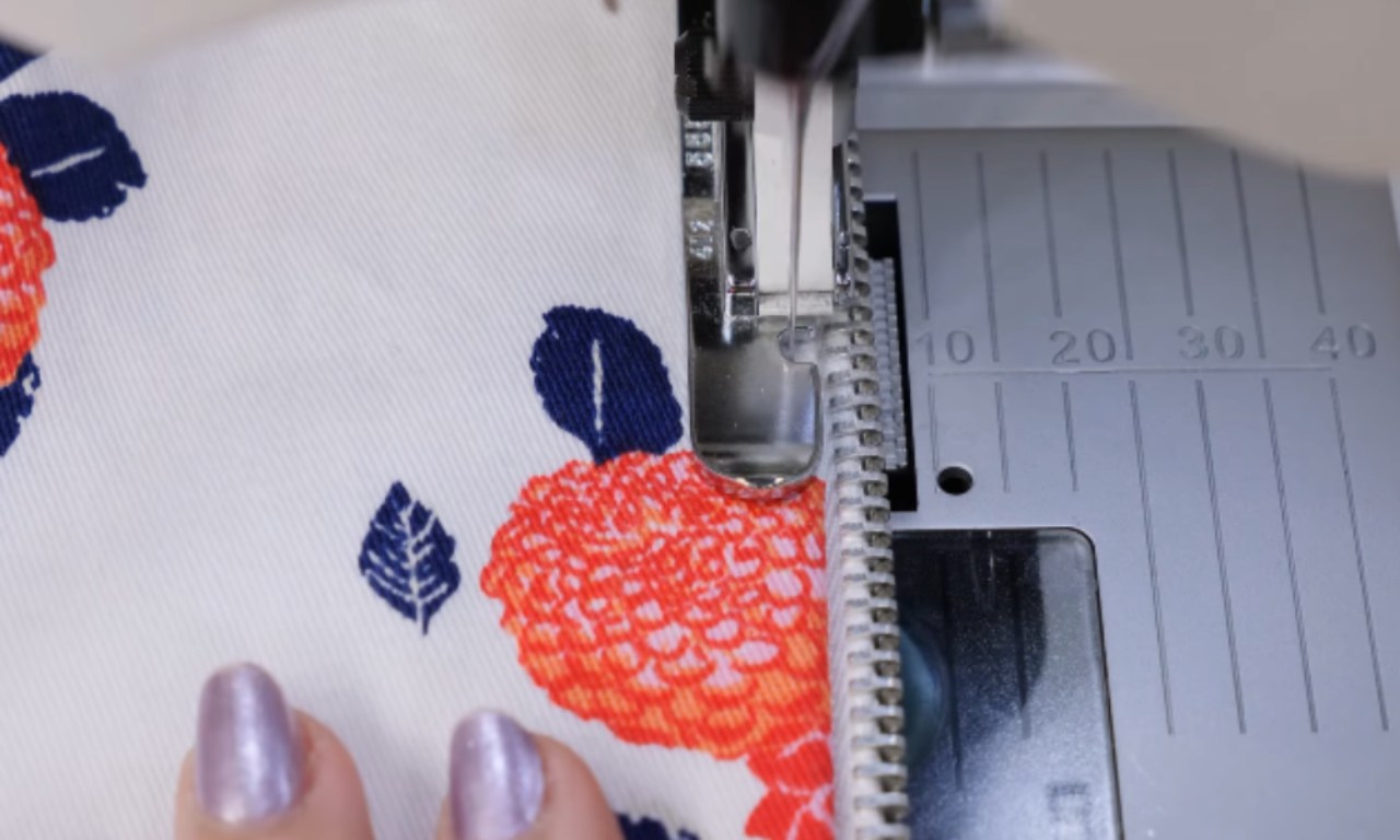 A close-up of a sewing machine stitching a zipper onto a piece of floral fabric.
