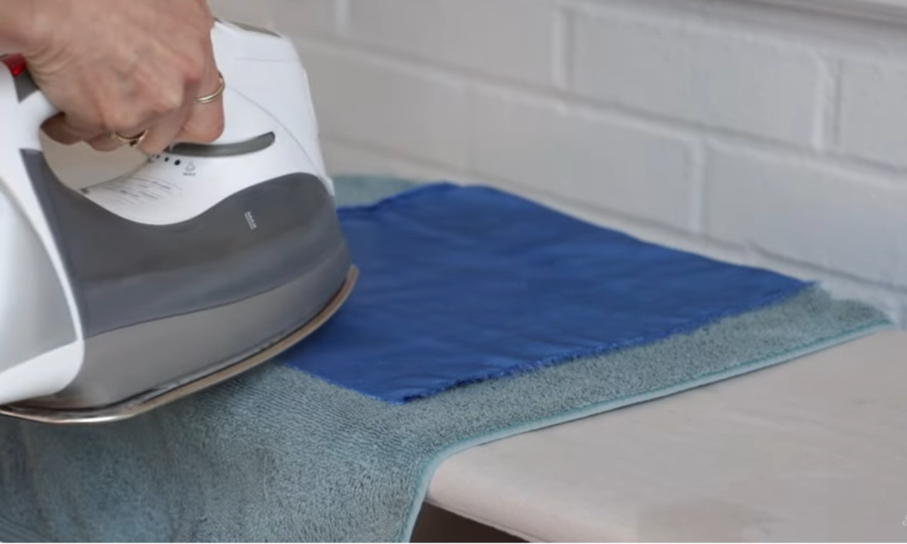 A person steaming a seam on blue velvet fabric by holding an iron just above it, with the fabric resting on a fluffy towel to protect the pile.