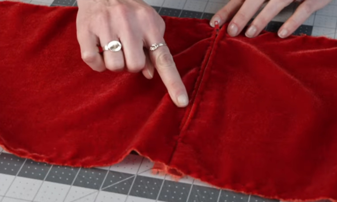 A person's hands pointing to a finished seam on a piece of red velvet fabric laid on a cutting mat, demonstrating a sewing technique.