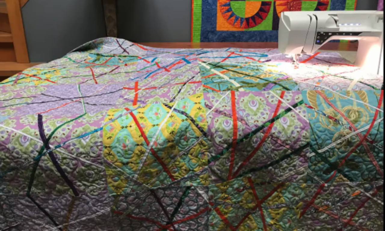 A large, colorful quilt made with fabric selvages and a dense quilting stitch, spread out in a sewing room with a sewing machine in the background.