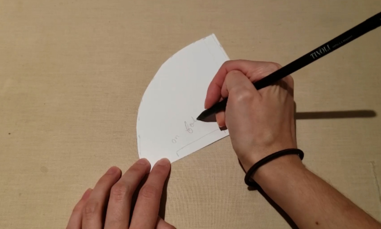 A person writing "on fold" with a black pencil on a white, quarter-circle paper pattern, which will be used to make a reusable coffee filter.