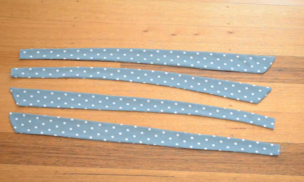 Four blue polka dot fabric strips for making bow tie shoulder straps, shown sewn into tubes and pressed flat on a wooden surface.