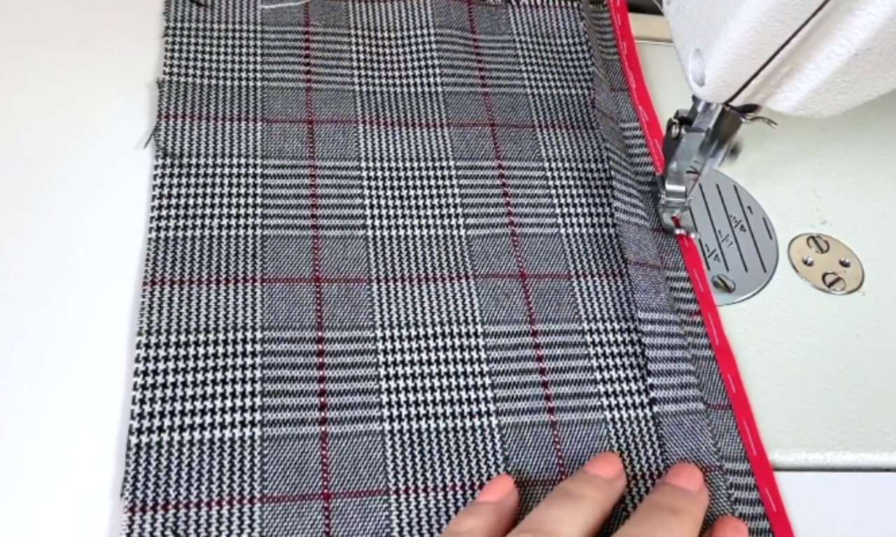 A close-up of a sewing machine stitching red bias tape to the raw edge of a black and white plaid fabric. A hand is visible, guiding the fabric.