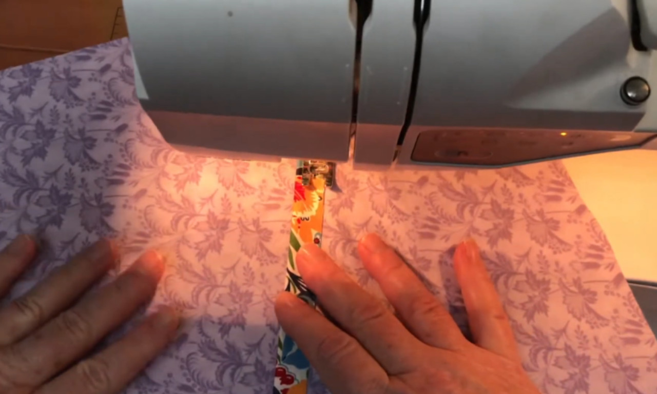 A close-up of a sewing machine stitching a colorful selvage strip onto purple floral fabric, with two hands guiding the material under the needle.