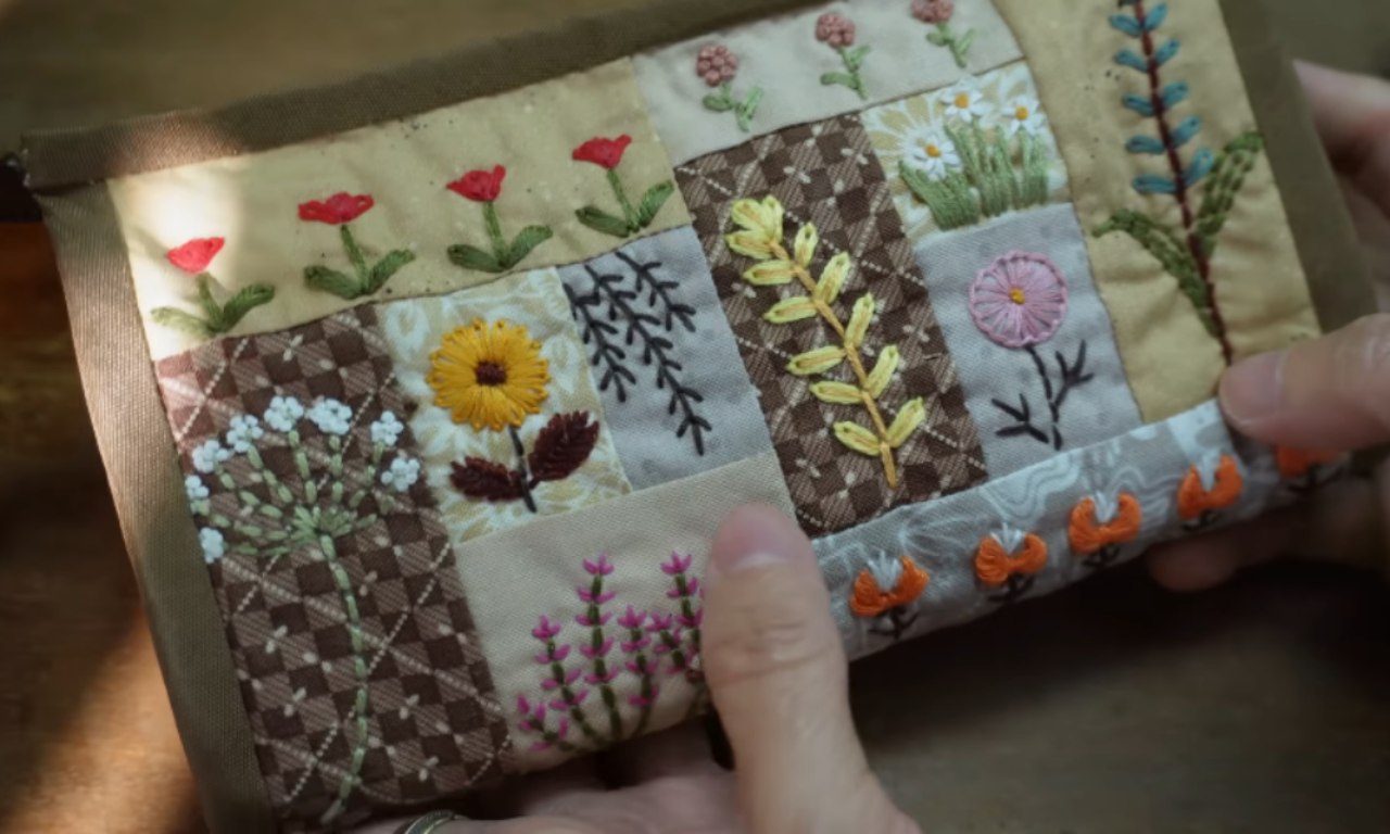 A close-up of a quilted pouch demonstrating embroidery on patchwork, with each fabric square featuring a different colorful, hand-stitched flower like a sunflower or daisy.