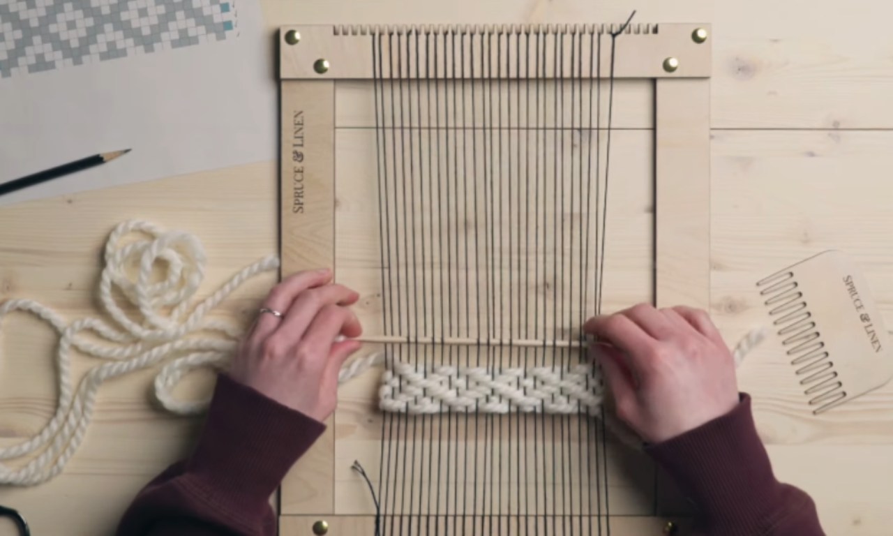 Overhead view of a person's hands weaving a braided pattern with thick white yarn onto a wooden frame loom with black warp threads. The loom is on a wooden table next to a weaving pattern, a pencil, and a wooden comb.