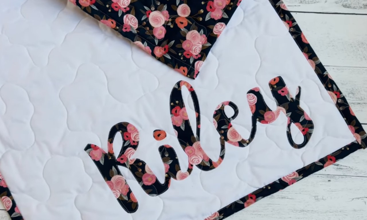 A finished personalized baby quilt showing appliqué letters that spell out the name "Riley" in a dark floral fabric on a white, stitched background.