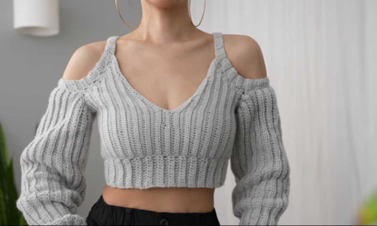 A woman models a light gray ribbed crochet crop top featuring long balloon sleeves and a cold-shoulder design with straps over the shoulders.