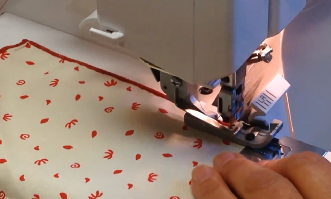 A serger sewing machine creating a narrow rolled hem with red thread on a piece of white patterned fabric as a hand guides it.