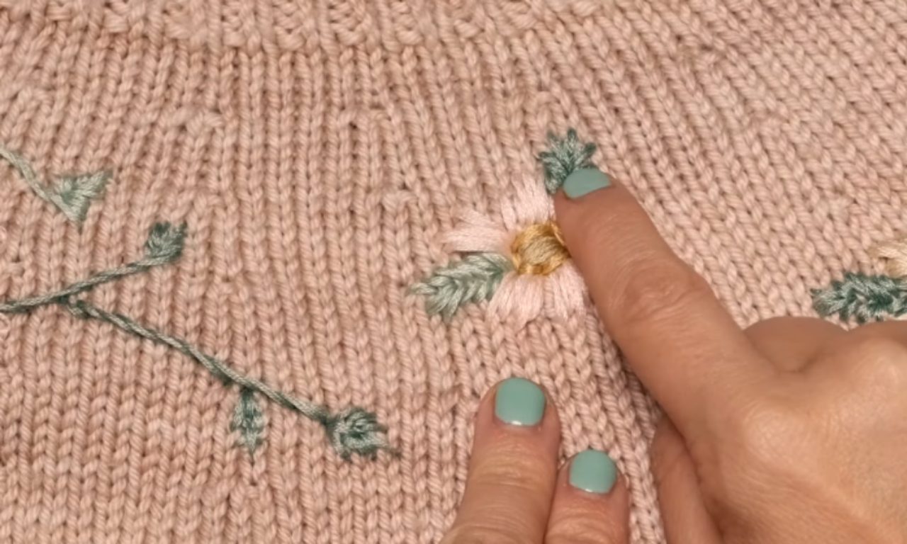 A close-up of a pink knit sweater with a hand-embroidered flower. A hand with mint green nail polish points to one of the flower's green leaves, showing the detail of the stitch.