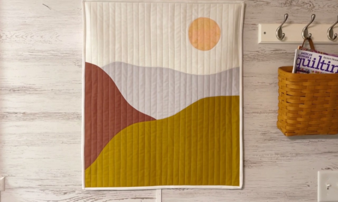 A modern landscape art quilt with an abstract sun and hills hanging on a white wood-paneled wall. To the right, a basket holding a quilting magazine hangs from a coat rack.