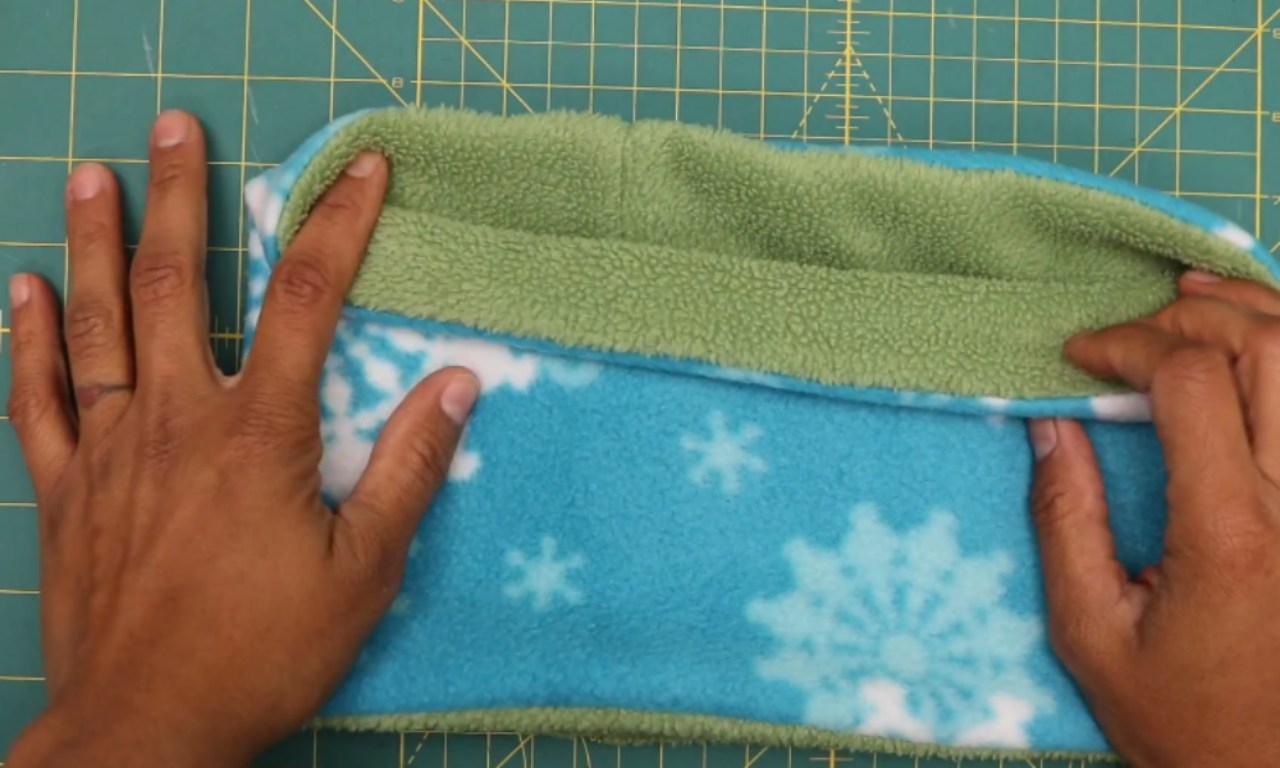 Hands folding a reversible fleece neck warmer, aligning the inner solid green fabric with the outer blue snowflake-print fabric on a green cutting mat.