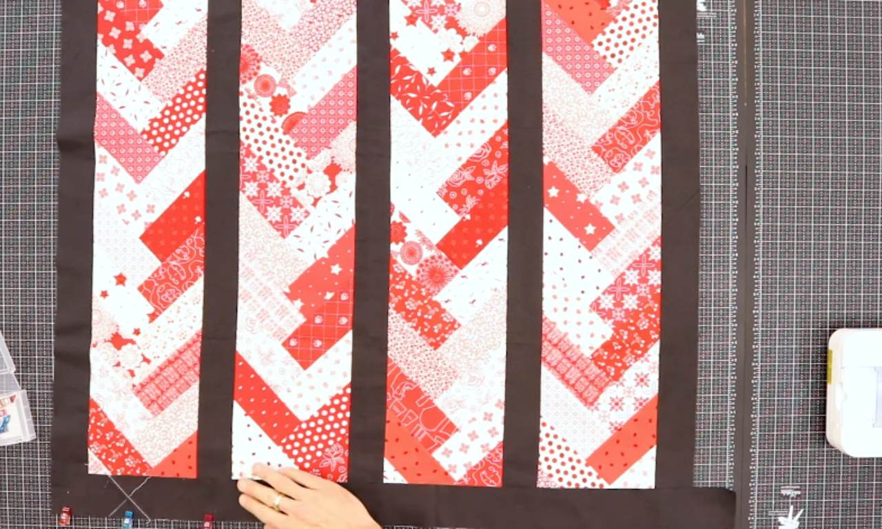 A nearly finished French braid quilt top with columns of red and white patterned fabrics, separated by black sashing, being laid out on a gridded cutting mat as a person attaches the final black border.