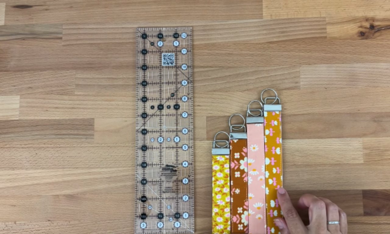 A collection of colorful, handmade fabric key fob wristlets with floral patterns lined up next to a quilting ruler on a wooden table, showing the different sizes possible for this DIY sewing project.