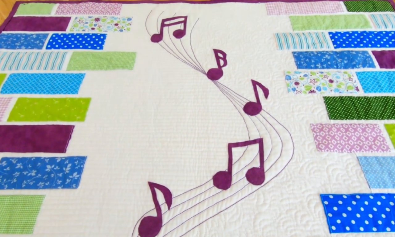 A finished music-themed quilt with a white center featuring appliquéd purple musical notes on a flowing, embroidered staff. The sides of the quilt are bordered with rows of colorful patterned fabric rectangles in shades of blue, green, and purple.