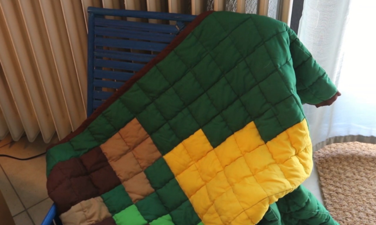 A finished DIY weighted blanket with a colorful, pixelated quilt design. The puffy squares of green, yellow, and brown fabric are draped over a blue chair indoors.