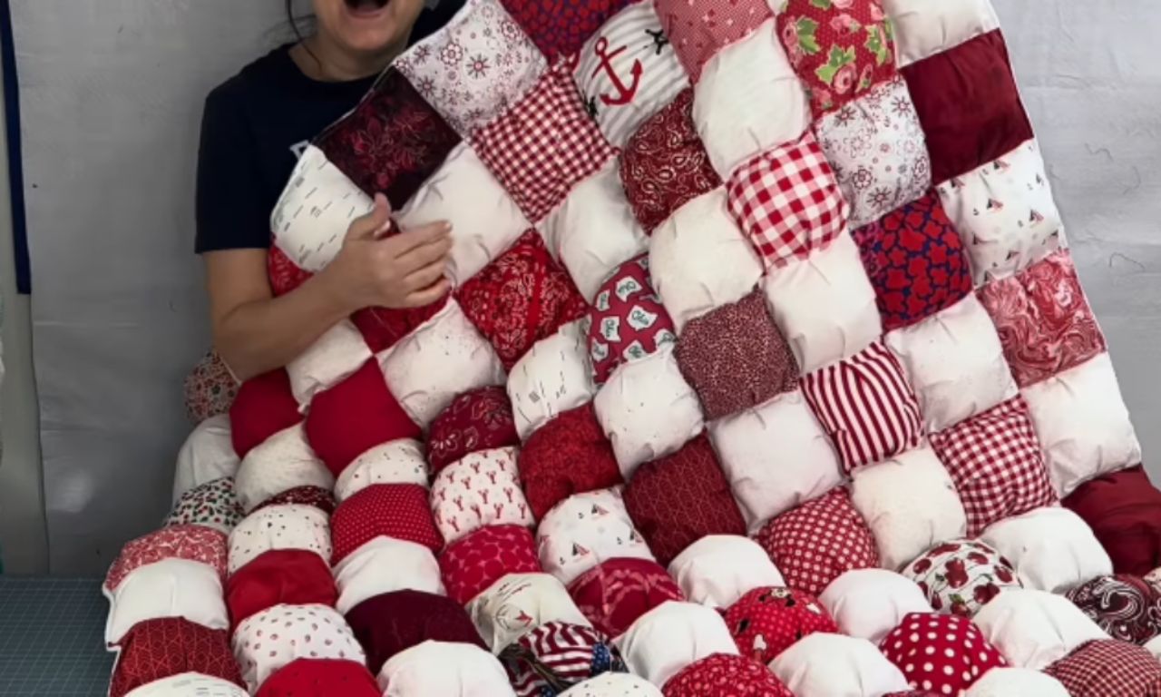 A woman joyfully holds up her finished red and white puff quilt, showing off the soft, 3D biscuit texture of the patterned squares.