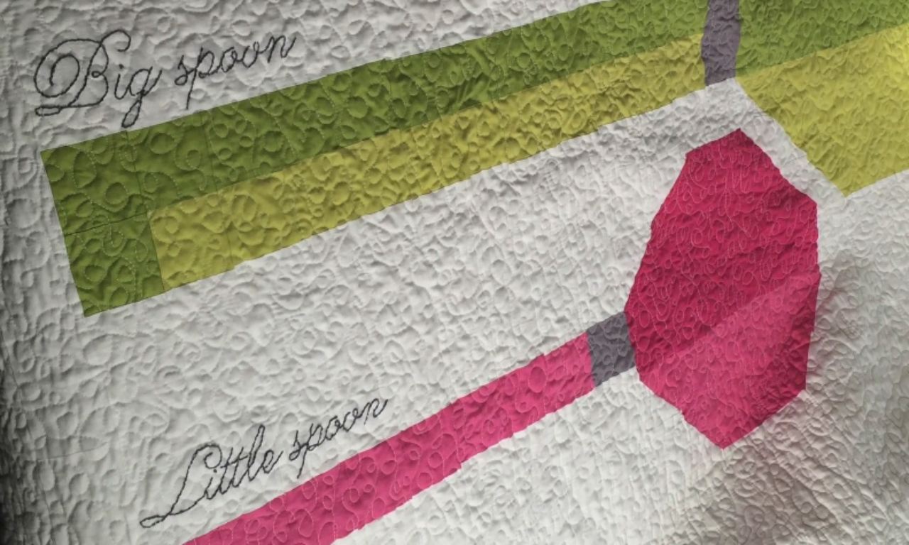 A close-up of a white quilt with a dense, swirly stitch pattern, featuring a modern, geometric design of two spoons with the words "Big spoon" and "Little spoon" embroidered in cursive.