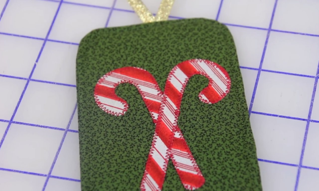 A close-up of a handmade fabric gift tag for Christmas, featuring two red-and-white striped candy canes appliquéd onto a green patterned background with a gold ribbon loop at the top.