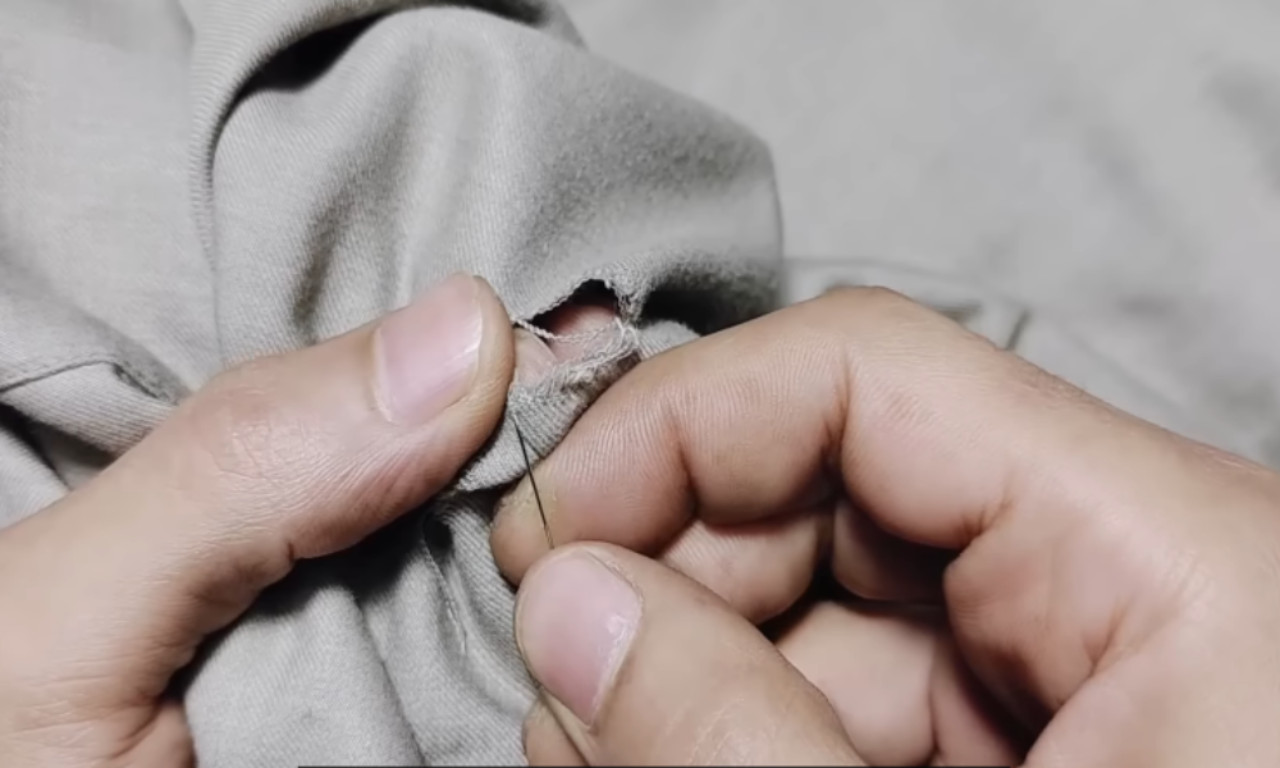 A person's hands carefully hand-sewing around a small hole in a grey shirt, reinforcing the fabric as the first step in a visible mending repair.