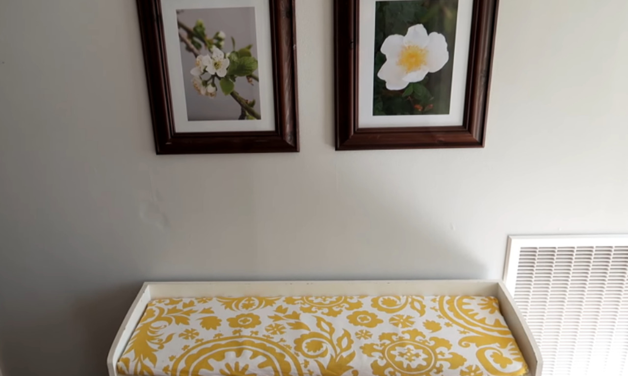 A finished DIY project showing a white bench with a custom-sewn bench seat cushion. The cushion features a bright yellow and white damask pattern and sits neatly inside the bench frame against a wall with two flower pictures.