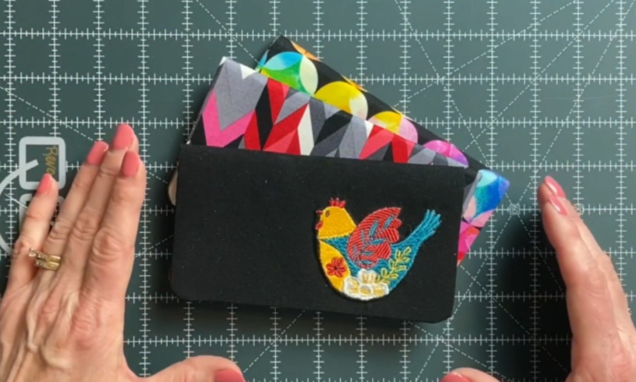 A crafter's hands displaying several finished DIY fabric checkbook covers on a green cutting mat. The main cover is black with a colorful embroidered bird, while two others behind it feature bright, geometric fabric patterns.