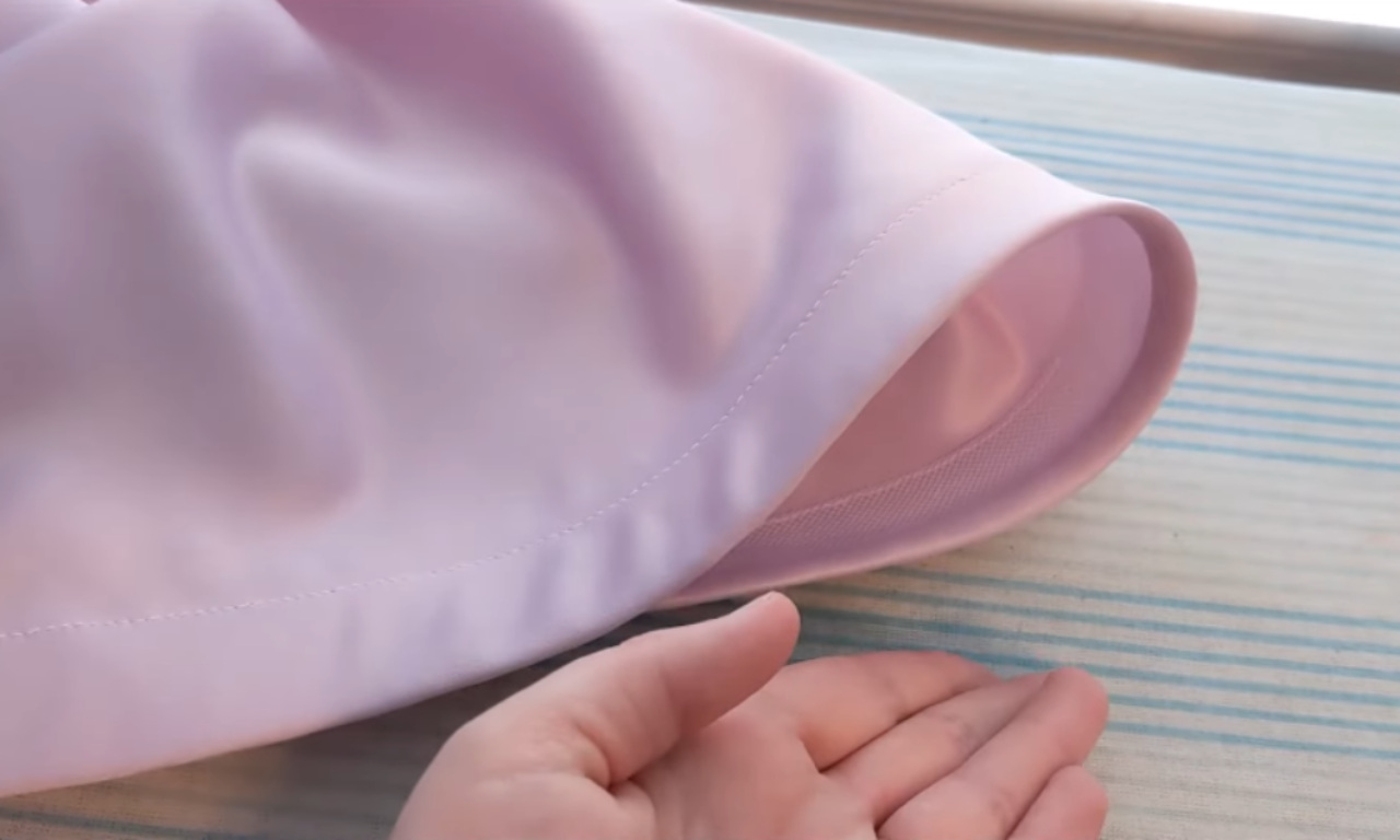 A hand presenting the finished hem of a light purple skirt, showing the clean topstitching on the outside and the white horsehair braid on the inside that creates a structured, wavy shape.