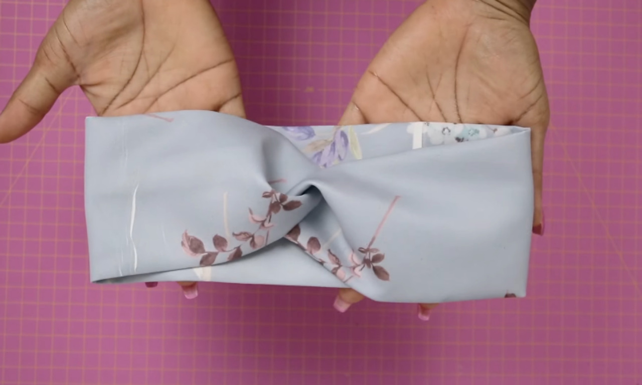 A pair of hands holding a finished light blue floral knot headband, showing the result of a DIY sewing tutorial against a pink cutting mat.