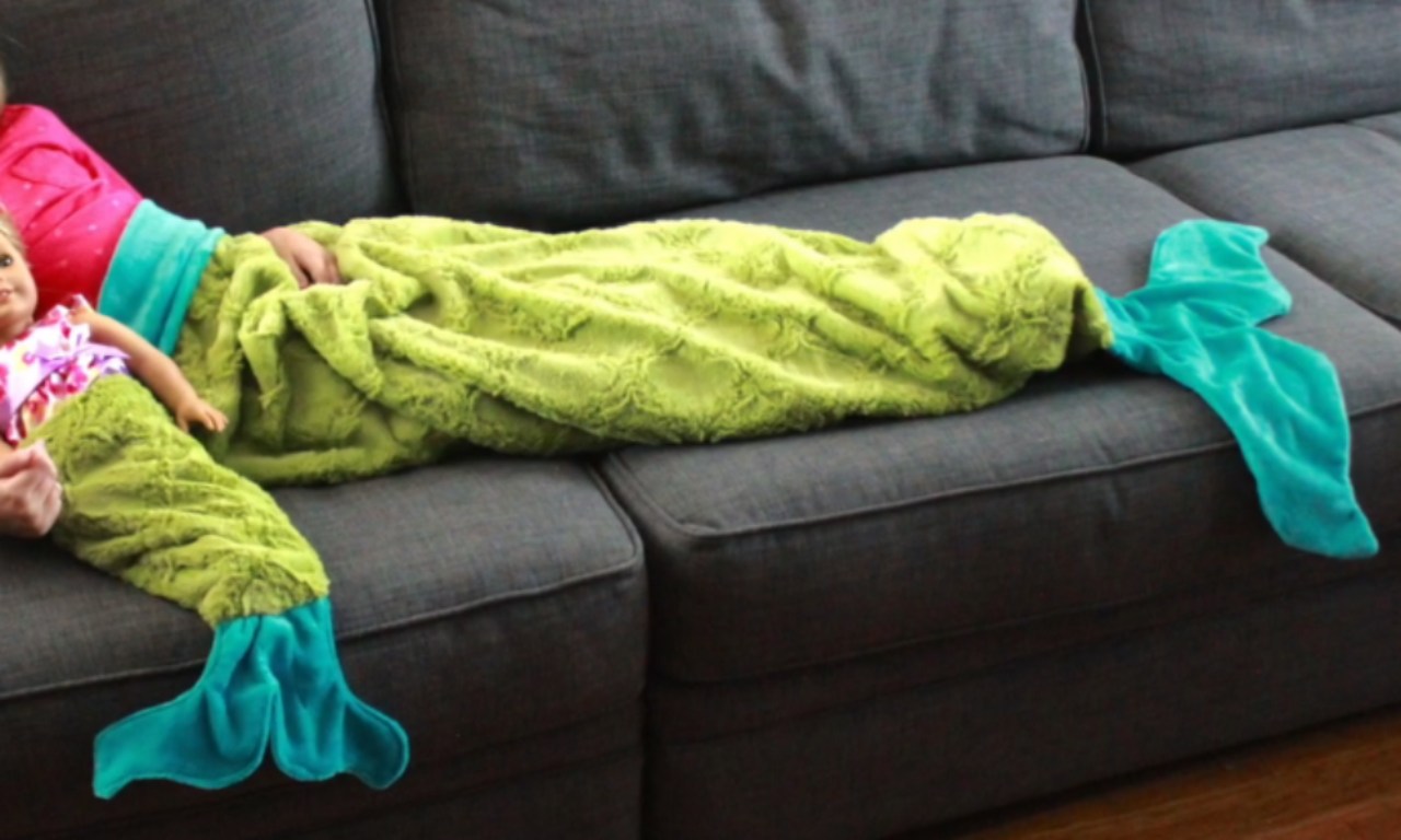 A child and a doll lie on a couch wearing matching, cozy lime green mermaid tail blankets with teal fins.